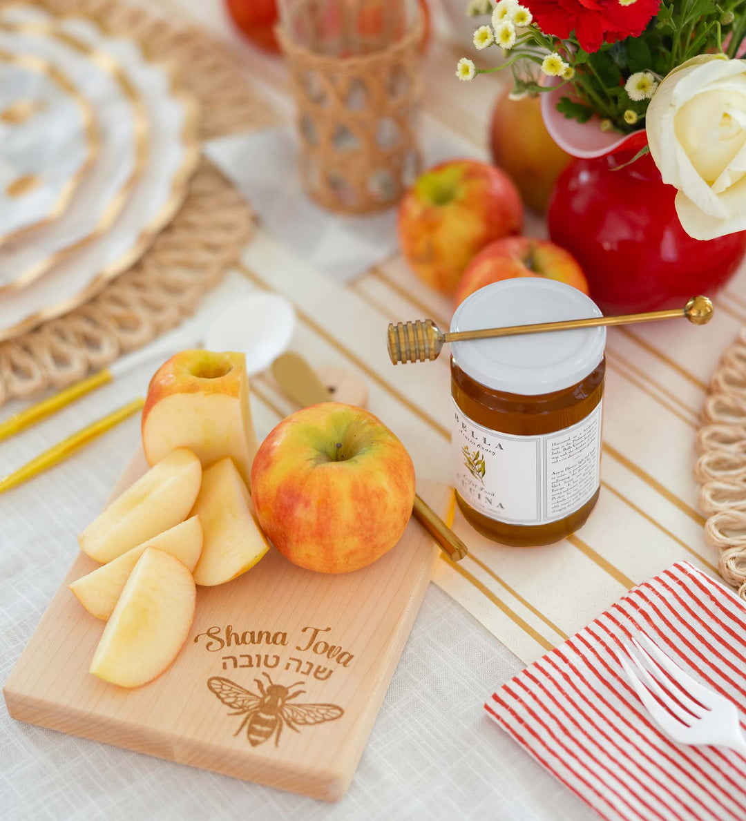 A cutting board with sliced apples, a whole apple, and “Shana Tova” engraved in Hebrew and English sits next to a honey jar filled with Bella Cucina Organic Thousand Flower Honey and a dipper, surrounded by flowers and red apples on a festive table setting.