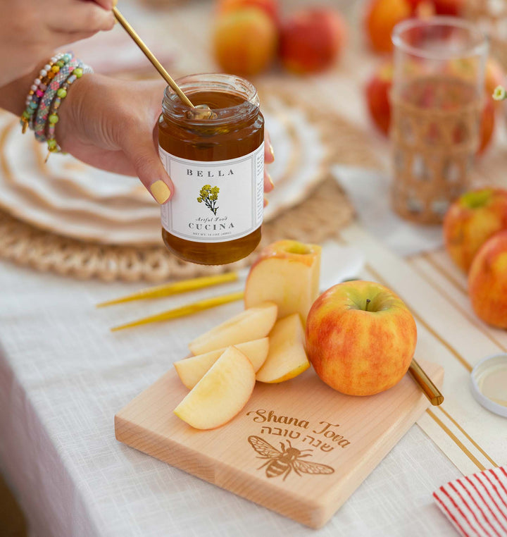 A hand dips a wooden stick into a jar of Bella Cucina Organic Thousand Flower Honey beside apple slices on a wooden board engraved with Shana Tova in English and Hebrew, and an apple, all arranged on a festive table setting.