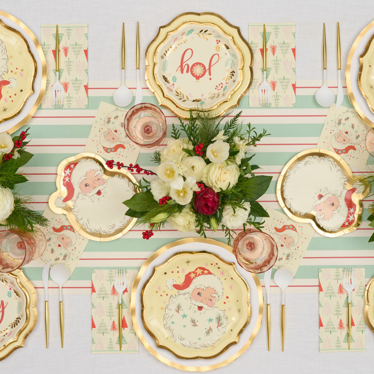 A Vintage Santa: Christmas Table for 16 tablescape featuring gold-rimmed disposable plates with Santa illustrations, tree-patterned napkins, gold-handled cutlery, and a floral centerpiece on a striped runner.