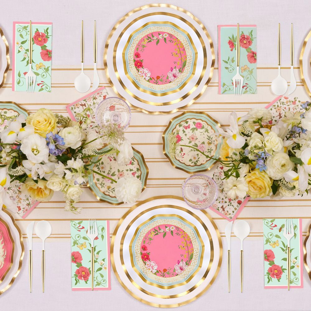 Elevate your event with the Ivory Rococo Table Setting by Sophistiplate—designer disposable plates with gold rims, paired with floral napkins, white cutlery, and lush flower centerpieces showcasing white and yellow blooms atop a light tablecloth—offering a sophisticated party-in-a-box experience.
