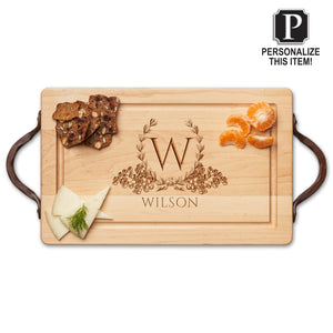 The Personalized Maple Wood Cutting + Cheeseboard by Maple features leather handles and is engraved with a floral wreath, the letter W, and the name Wilson. It holds crackers, cheese, orange segments, and displays a "Personalize This Item!" label.