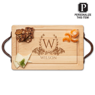 The Personalized Maple Wood Cutting + Cheeseboard by Maple features leather handles and is engraved with a floral wreath, the letter W, and the name Wilson. It holds crackers, cheese, orange segments, and displays a "Personalize This Item!" label.