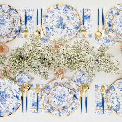 Create a luxe party-in-a-box with the Timeless Table Setting by Sophistiplate—featuring designer blue and white floral plates, gold utensils, blue-handled cutlery, and baby's breath centerpiece on a white tablecloth.